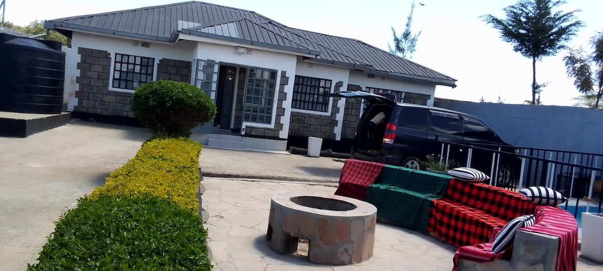 The exterior of a single-story house is showcased, featuring a gray metal roof and a stone facade. A circular fire pit is positioned in a paved seating area, surrounded by colorful outdoor seating. Lush greenery borders the pathway leading to the entrance, with a parked vehicle nearby.