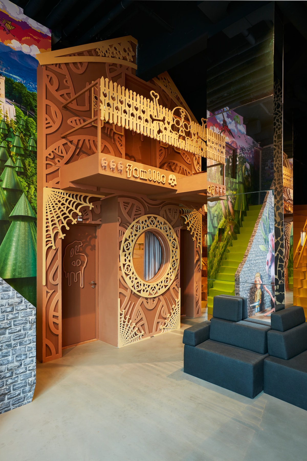An artistic entry features an intricately designed brown facade, combining natural motifs with elaborate patterns. A circular mirror is set within the door frame, which is accentuated by a carved balcony above. The vibrant mural background enhances the whimsical ambiance of the space.