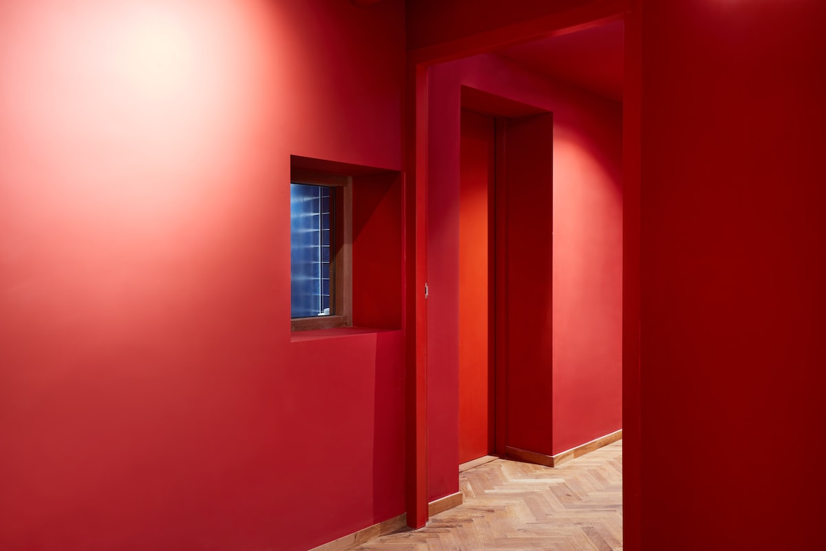 A vibrant red hallway is highlighted by a small window and a doorway leading to another space. The wooden flooring features a herringbone pattern, complementing the warm tones of the walls. Light is softly filtered through the window, creating a dynamic contrast.