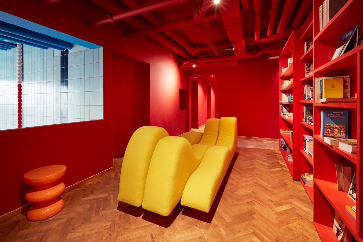 A vibrant lounge area is featured, characterized by bold red walls and a unique yellow wavy sofa. A small orange side table complements the seating. Open shelving displays an array of books, enhancing the creative ambiance of the space.