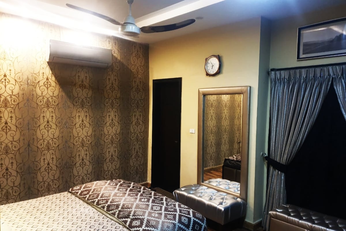 A stylish bedroom features patterned wall decor and a comfortable bed with an elegant throw. A large mirror reflects the space, while a clock is mounted above the door. Soft lighting is provided by an overhead fan and wall-mounted air conditioning unit.