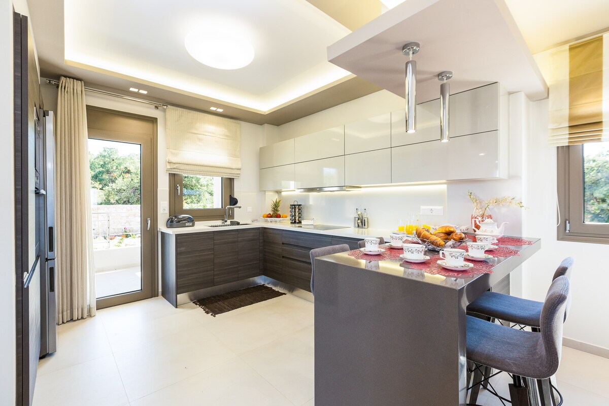 A modern kitchen is visible, featuring sleek cabinetry and a large island with bar seating. Natural light enters through a window, illuminating the space. High-end appliances are integrated into the design, and a table is set with dishes, suggesting a welcoming atmosphere for meals.