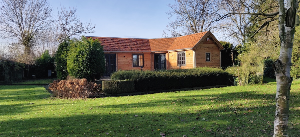 A charming wooden chalet is surrounded by a lush green garden, featuring well-maintained hedges and trees. The exterior showcases a sloped roof and multiple windows for natural light. A peaceful setting offers ample outdoor space for relaxation.