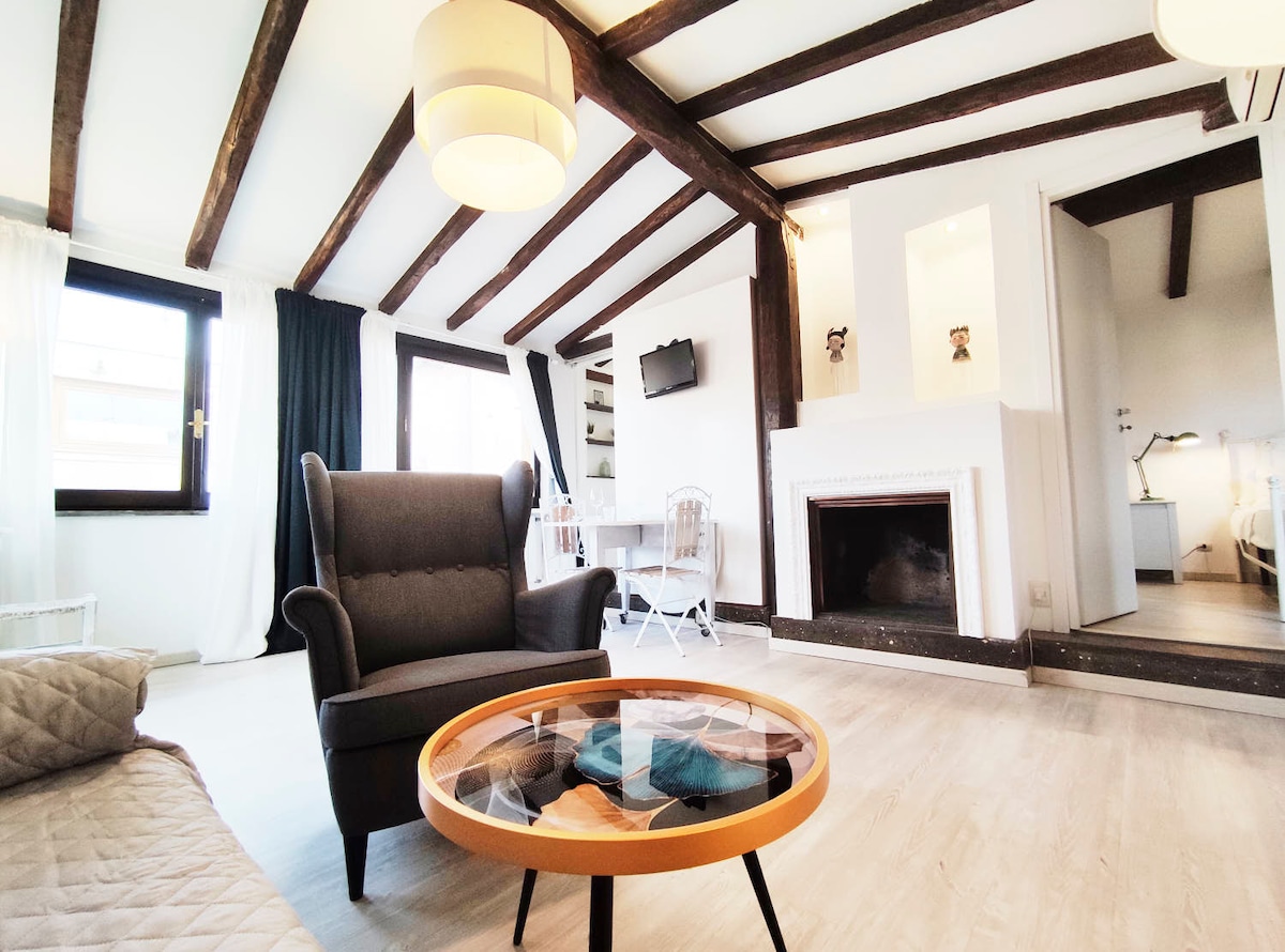 A spacious living room is accented by wooden beams and a modern ceiling light. A comfortable armchair and a circular coffee table are situated in the center. The room features large windows allowing natural light to fill the space, and a flat-screen TV is mounted on the wall.