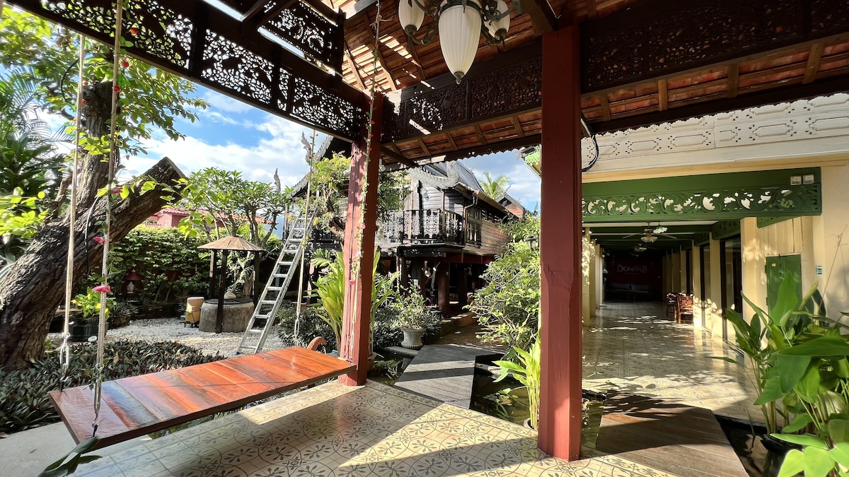 A welcoming porch features a wooden swing and intricate wooden detailing, surrounded by lush greenery. A serene pathway leads to a decorative courtyard, while sunlight streams through overhead structures, illuminating the vibrant terrace and adjoining spaces.