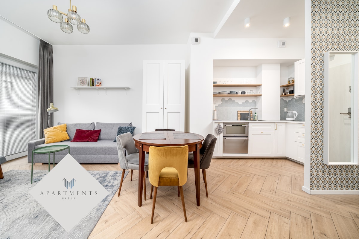 A modern open-concept living area is presented, featuring a light gray sofa with colorful accent cushions and a round wooden dining table surrounded by upholstered chairs. A stylish kitchen area with white cabinets and sleek appliances is visible, complemented by natural light from large windows.