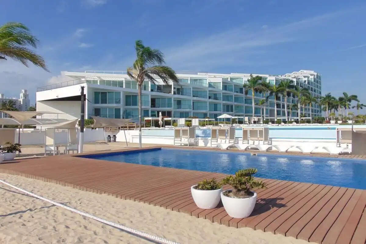 A spacious outdoor area features a large swimming pool surrounded by wooden decking. Comfortable lounge chairs are positioned near the pool, with tropical palm trees and a modern condominium building visible in the background.