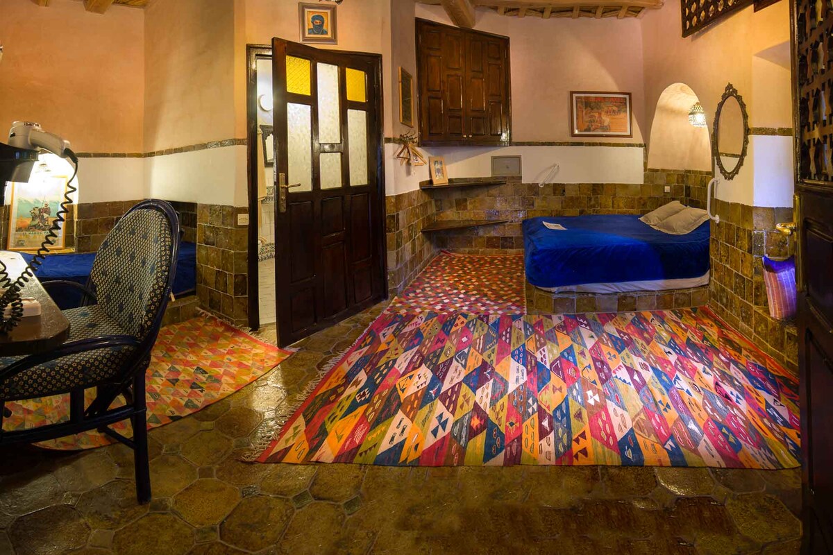 A cozy interior is showcased with textured stone walls and a colorful patterned rug. A spacious bed with blue bedding is positioned alongside a compact seating area featuring a table and a chair. Light enters through the door and windows, illuminating the warm tones of the space.