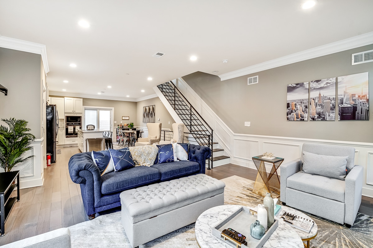 The open-concept living area features a blue sectional sofa and light gray armchairs, set against a backdrop of neutral walls. A round marble coffee table sits atop a textured area rug, while artwork adorns the wall. Natural light fills the space, enhancing the inviting atmosphere.