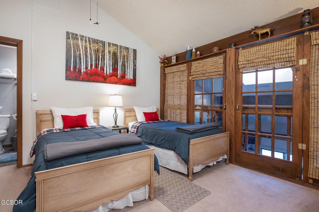 A well-lit bedroom features two twin beds with dark blue bedding, neatly arranged beside a bedside lamp. A wooden accent wall displays nature-themed decor. The room includes a door leading to a private balcony, and a bathroom is visible through an open door on the left.