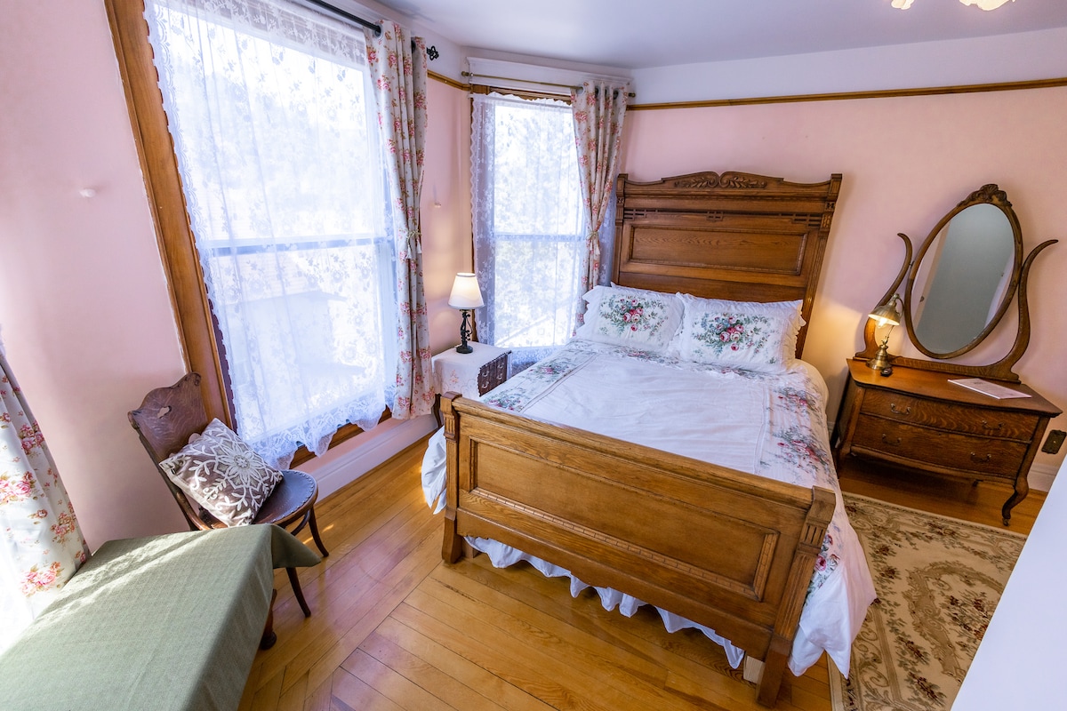 The room features a queen-size sleigh bed adorned with floral bedding, complemented by antique furnishings. Two large windows framed with lace curtains allow natural light to fill the space. A small chair and table are positioned near the window, enhancing the room's charm.