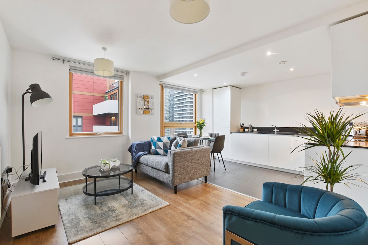 A modern living area is displayed, featuring a comfortable sofa with cushions and a glass coffee table. Natural light enters through large windows, illuminating a fully equipped kitchen in the background. A potted plant adds a touch of greenery to the space.