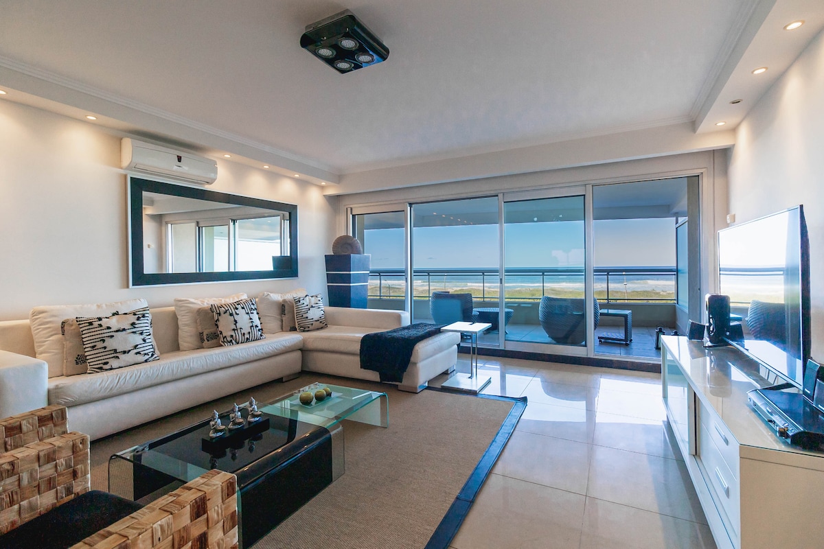 A modern living area showcases a spacious white sofa adorned with patterned cushions. Large sliding glass doors offer unobstructed ocean views, while a sleek television is positioned on a minimalistic white cabinet. Warm lighting enhances the contemporary decor and ambiance.