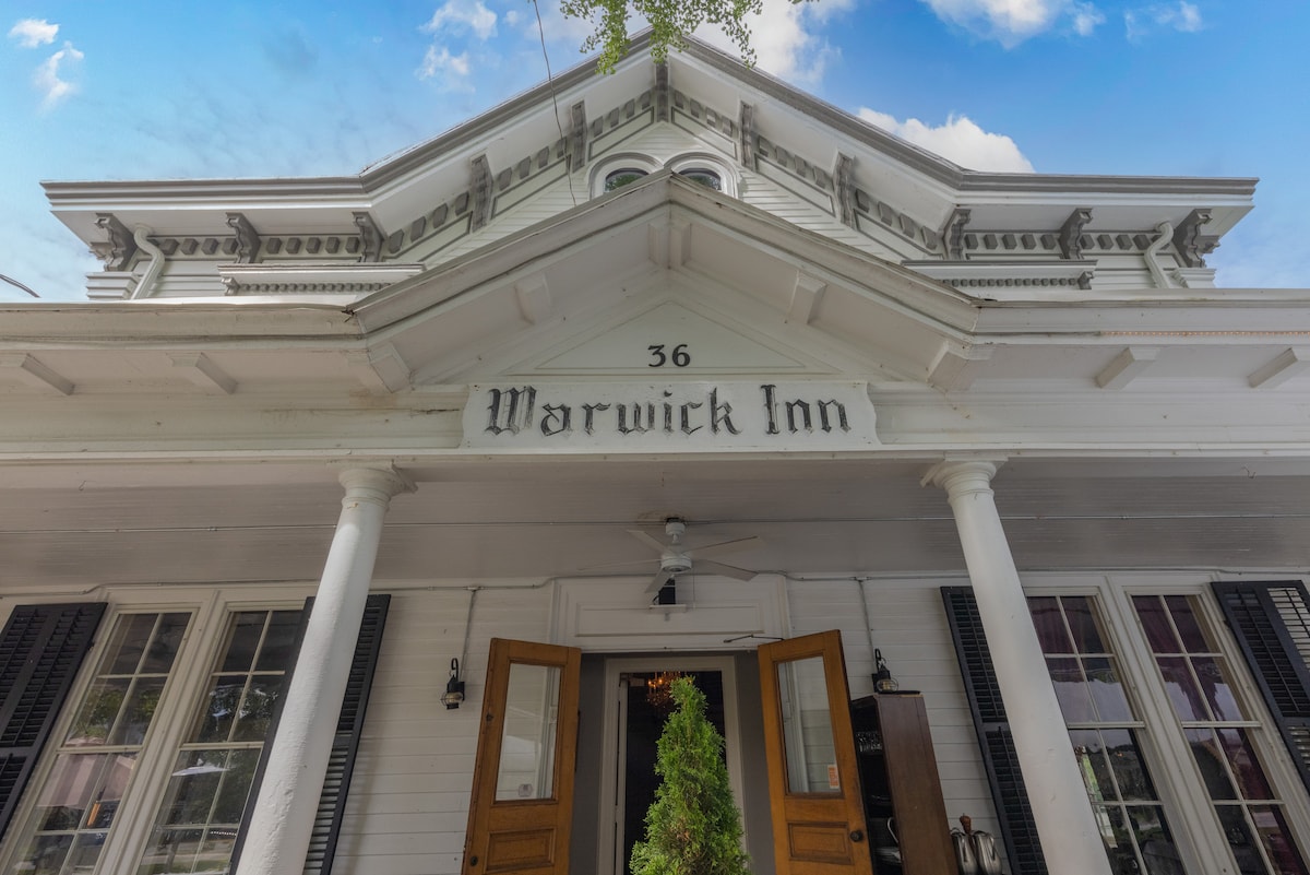 The exterior of the Warwick Inn is showcased, featuring classic white siding and intricate architectural details. A central entrance with double doors is framed by dark shutters, while the building's name is elegantly displayed above. Lush greenery is positioned near the entrance, adding to the inviting appearance.