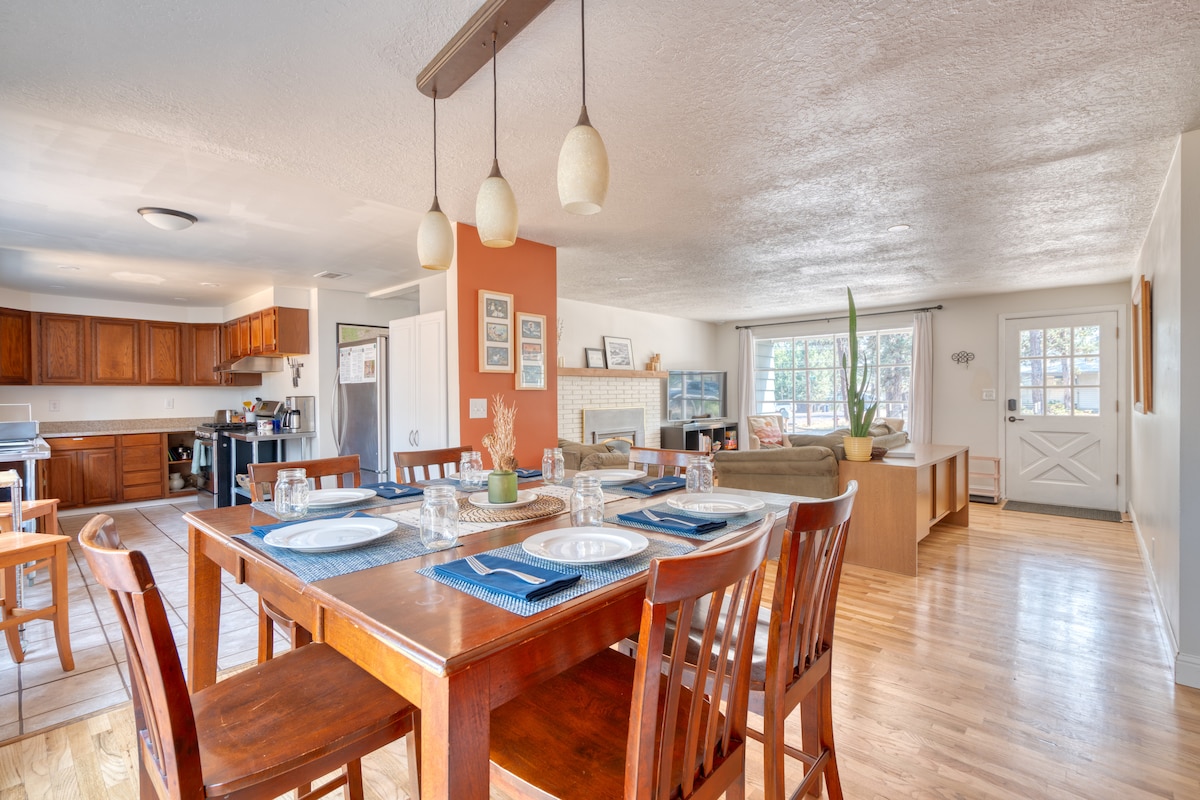 A spacious dining area is highlighted by a wooden table set with plates and glassware, surrounded by wooden chairs. The open layout allows visibility into the inviting kitchen and living area, where natural light fills the room through large windows.