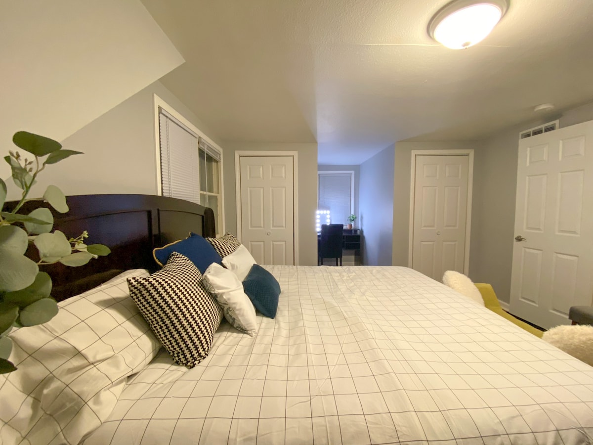 A spacious bedroom is furnished with a neatly made bed featuring a grid-patterned duvet and various accent pillows. Light-colored walls are complemented by a workspace visible through open doors, enhancing functionality within the room.