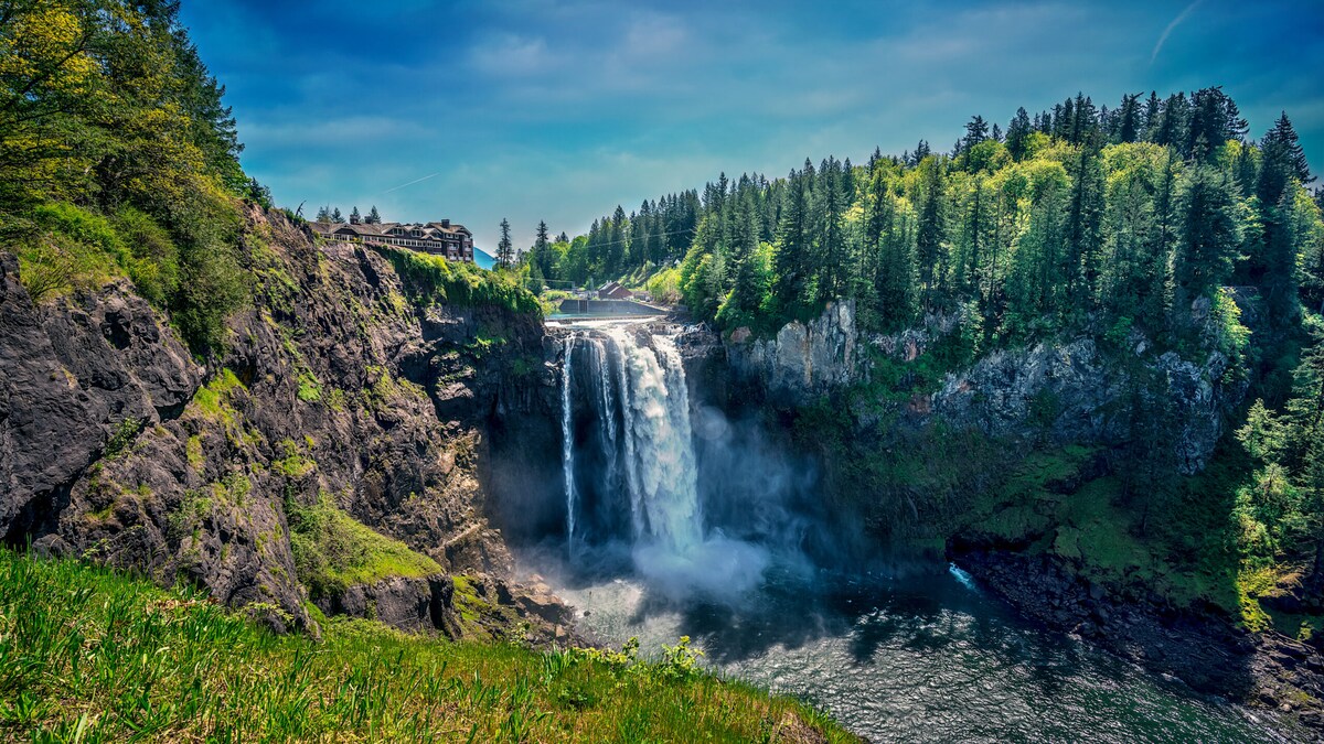 A cascading waterfall is observed flowing into a tranquil body of water, surrounded by lush greenery and rocky cliffs. The vibrant sky adds a bright backdrop to the natural landscape, highlighting the serene beauty of the area.