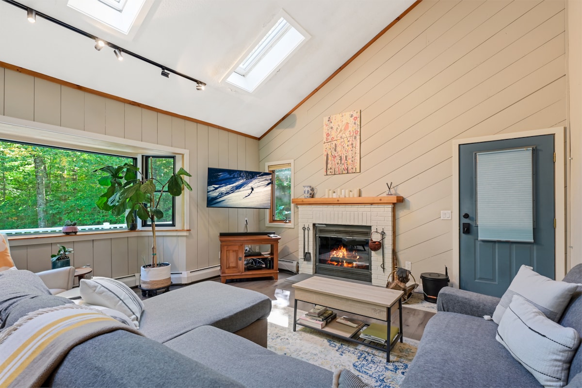 A bright living area features a large sectional sofa and a cozy fireplace, drawing attention to the warm light from the skylights. A television is mounted above the fireplace, and greenery from a nearby plant enhances the inviting setting. Windows provide views of the surrounding trees.