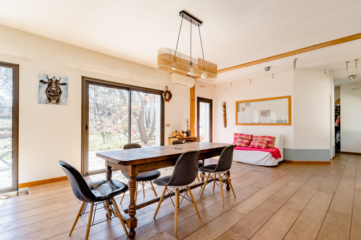 A dining area features a large wooden table surrounded by stylish chairs. Large sliding glass doors allow natural light and offer views of the exterior garden. A comfortable seating area is visible in the background, along with minimalistic decor on the walls.