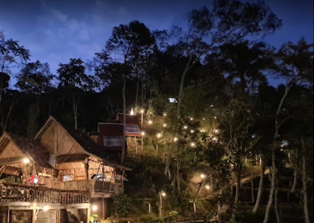 Two rustic wooden buildings are visible against a backdrop of trees. Soft lights adorn the path leading up the hillside, providing a warm glow in the evening setting. The surrounding nature creates a serene and inviting ambiance.
