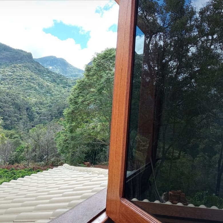 A wooden window frame reflects lush greenery and mountain views. The scene captures the natural beauty surrounding the space, with patches of blue sky and white clouds visible beyond the foliage.