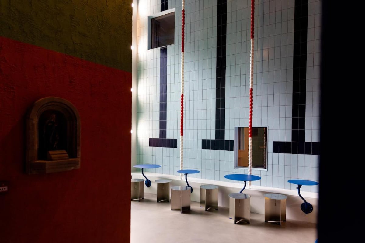 A spacious room showcases vibrant turquoise tiles and walls painted in green and red. Blue tables are positioned against two opposite walls, complemented by hanging ropes. Natural light filters through large windows, highlighting the unique color palette and design elements.