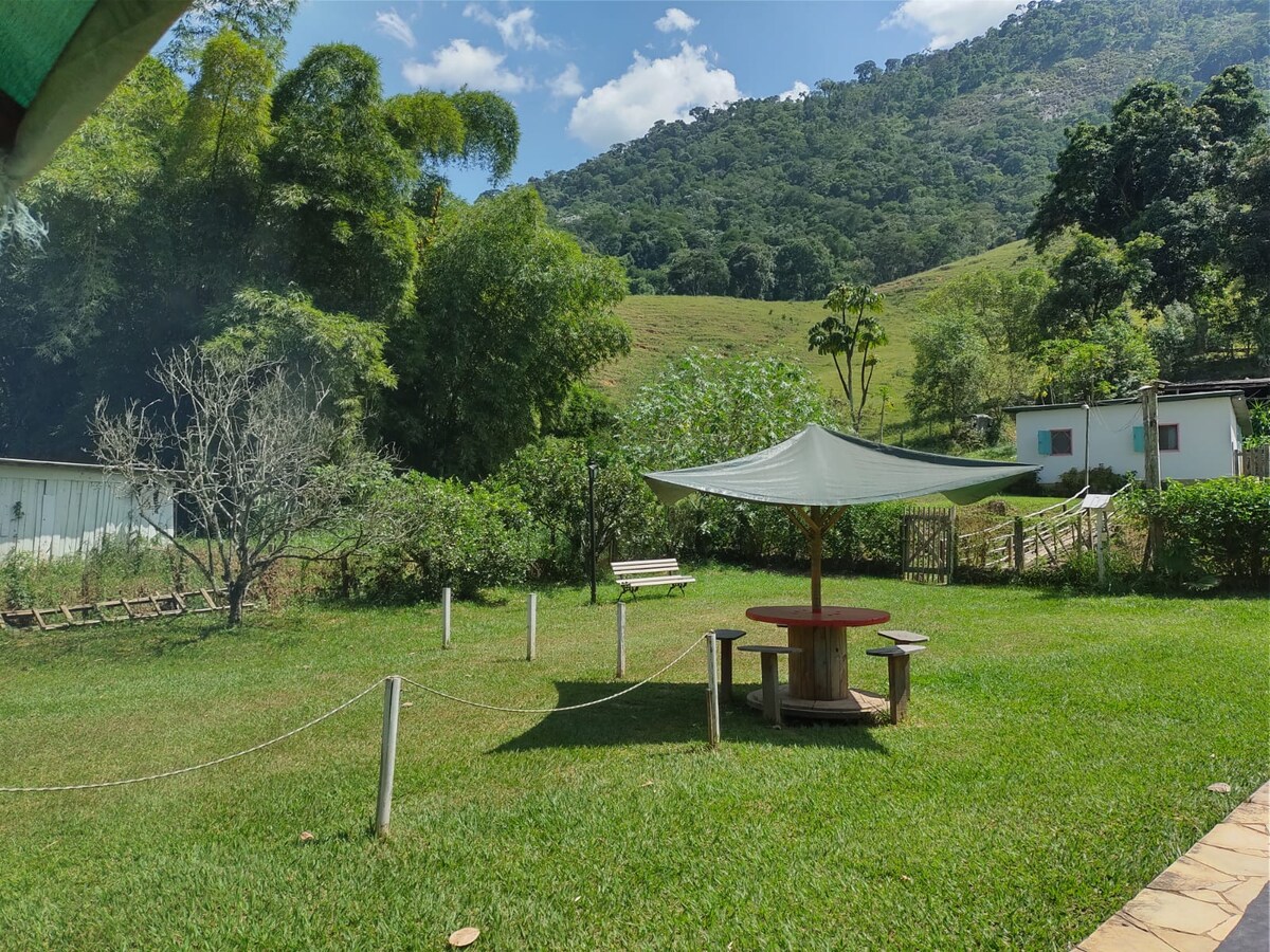 A grassy outdoor area is featured, with a shaded picnic table at its center. Surrounding greenery includes trees and plants, while a hillside is visible in the background, complemented by scattered clouds in a bright blue sky.