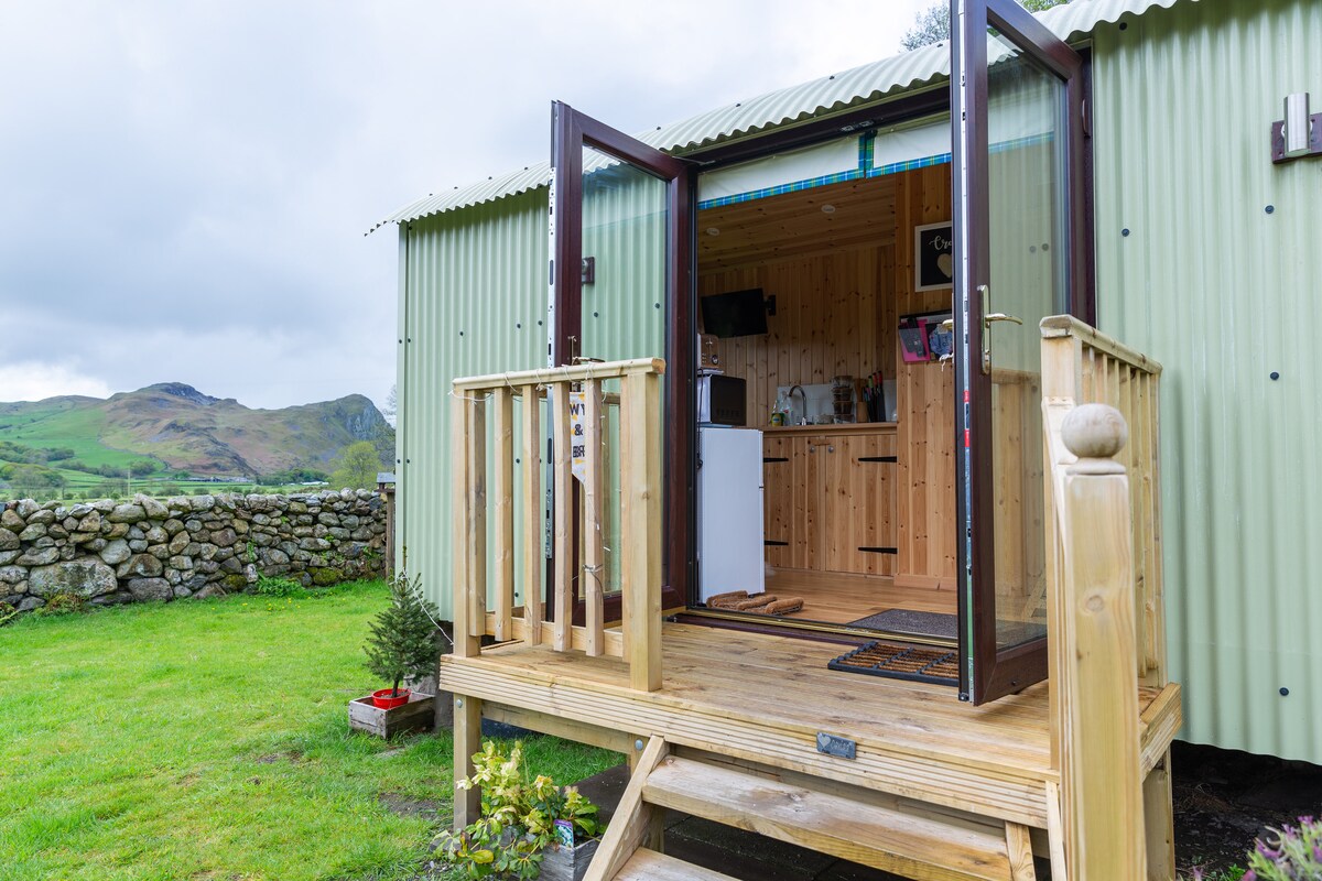 The exterior of the Shepherd's Hut showcases a welcoming entrance with double doors open to reveal a cozy interior. A wooden deck leads to grassy surroundings, complemented by a stone wall and rolling hills in the background.