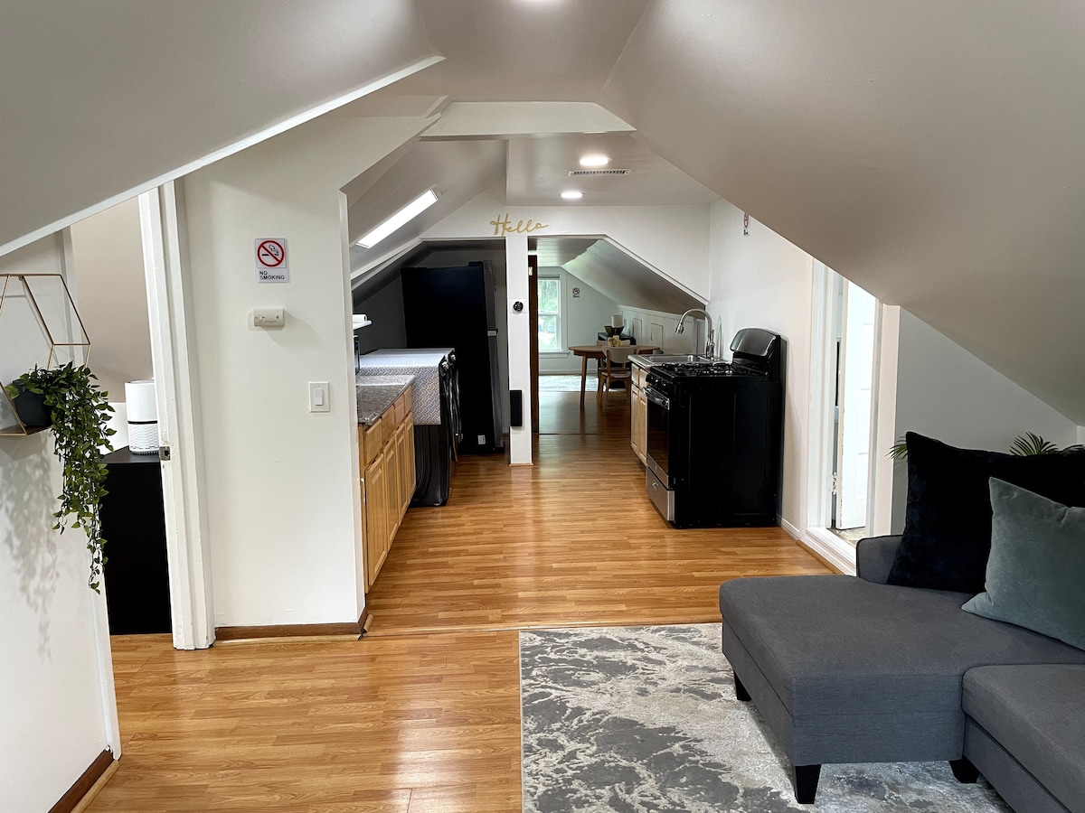 The image displays an open living area with wood flooring and sloped ceilings. A dark sofa is positioned beside a light rug. The kitchen area features wooden cabinets and black appliances, while a dining table is visible in the distance, inviting natural light through a nearby door.