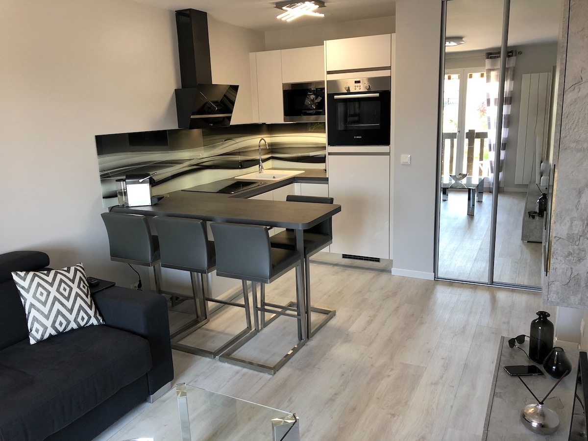 The kitchen area features modern cabinetry and an island with four gray stools. A sleek black backsplash complements the culinary space, which includes appliances seamlessly integrated into the design. Light-colored flooring enhances the open atmosphere of the living area, with a comfortable couch positioned nearby.