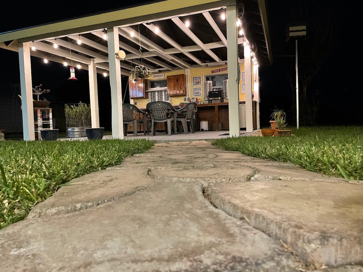 A well-lit outdoor kitchen is featured under a covered patio, with multiple chairs gathered around a table. A stone pathway leads to the space, bordered by lush green grass and potted plants, creating a welcoming area for gatherings.