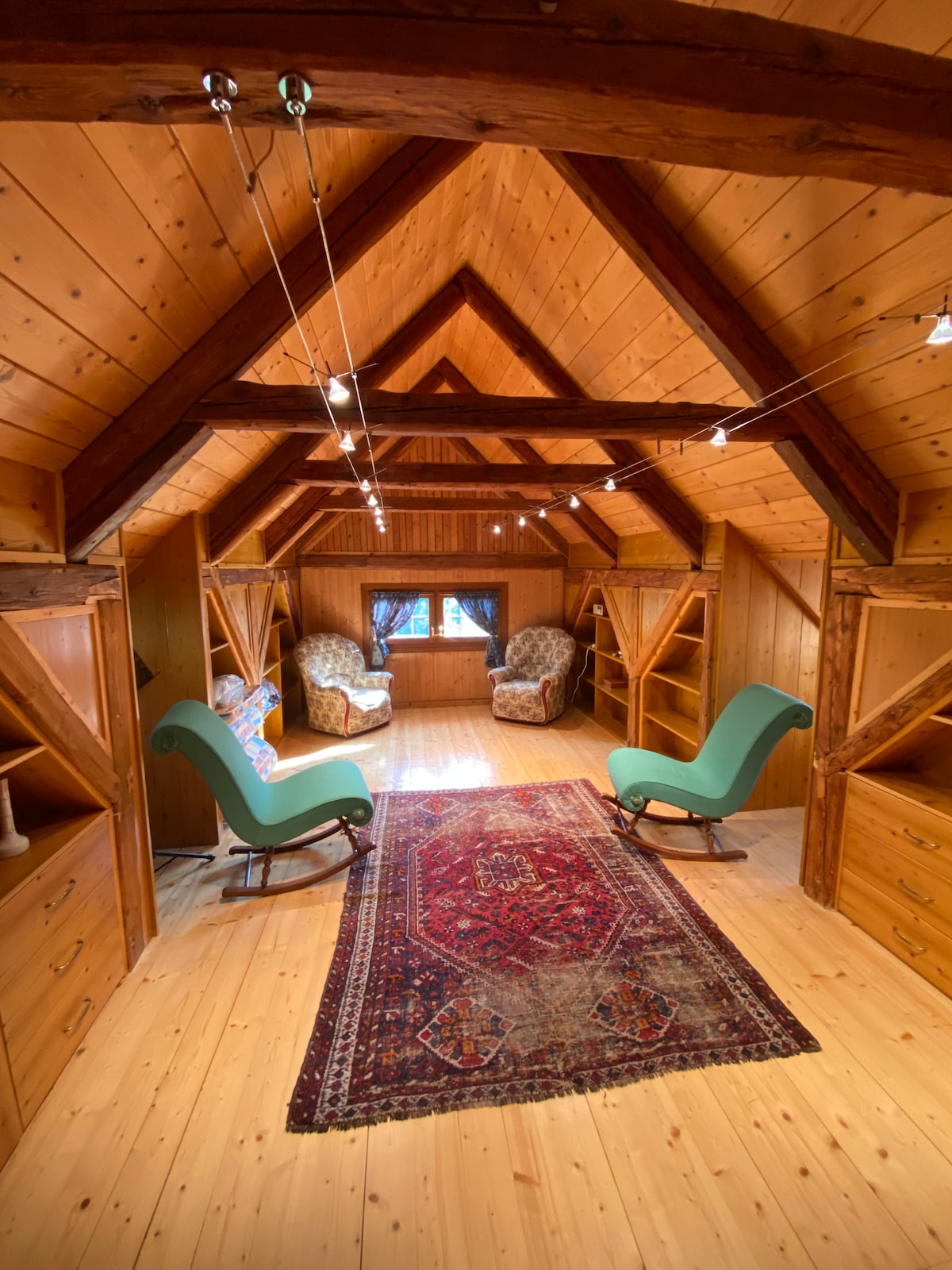 A wooden interior features vaulted ceilings with exposed beams and a warm wooden floor. A colorful area rug lays in the center, while two comfortable, patterned armchairs are positioned beside a window. Built-in shelves line the walls, adding to the cozy ambiance.