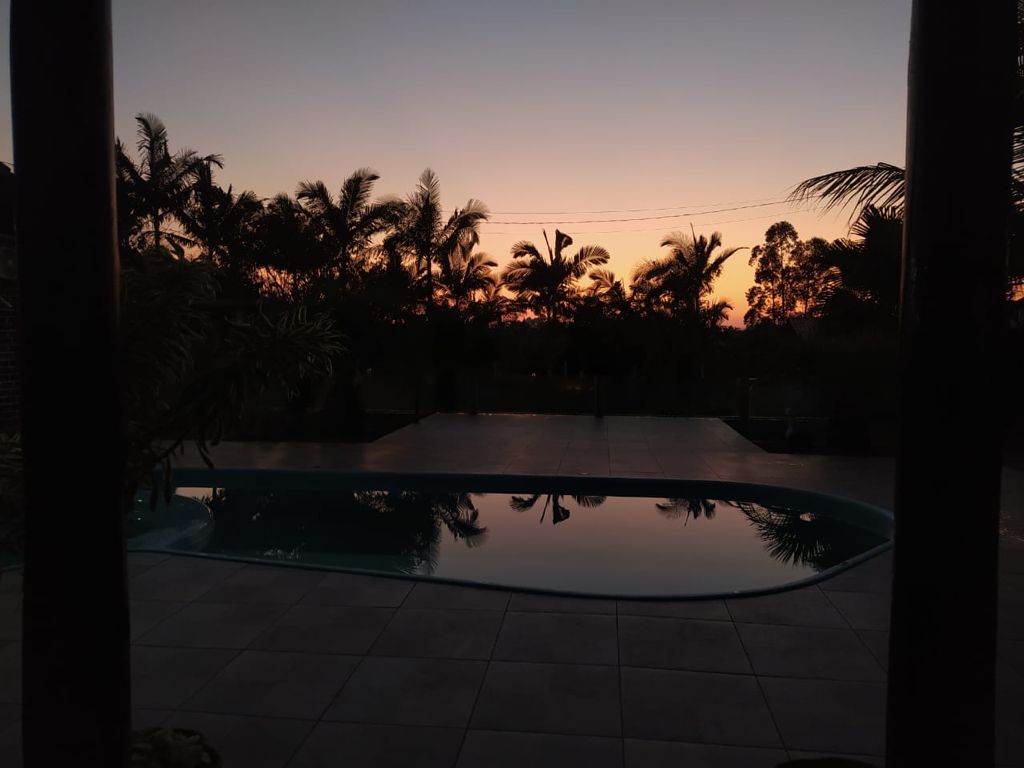 A peaceful view of a swimming pool at sunset is captured, with the vibrant colors of the sky reflecting in the calm water. Silhouetted palm trees frame the scene, creating a serene atmosphere that invites relaxation.