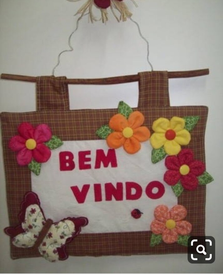 A welcoming sign is prominently displayed featuring the words 'Bem Vindo' in bold red letters. The sign is adorned with colorful fabric flowers in shades of orange, pink, and green, alongside a stylized butterfly, adding a cheerful touch.