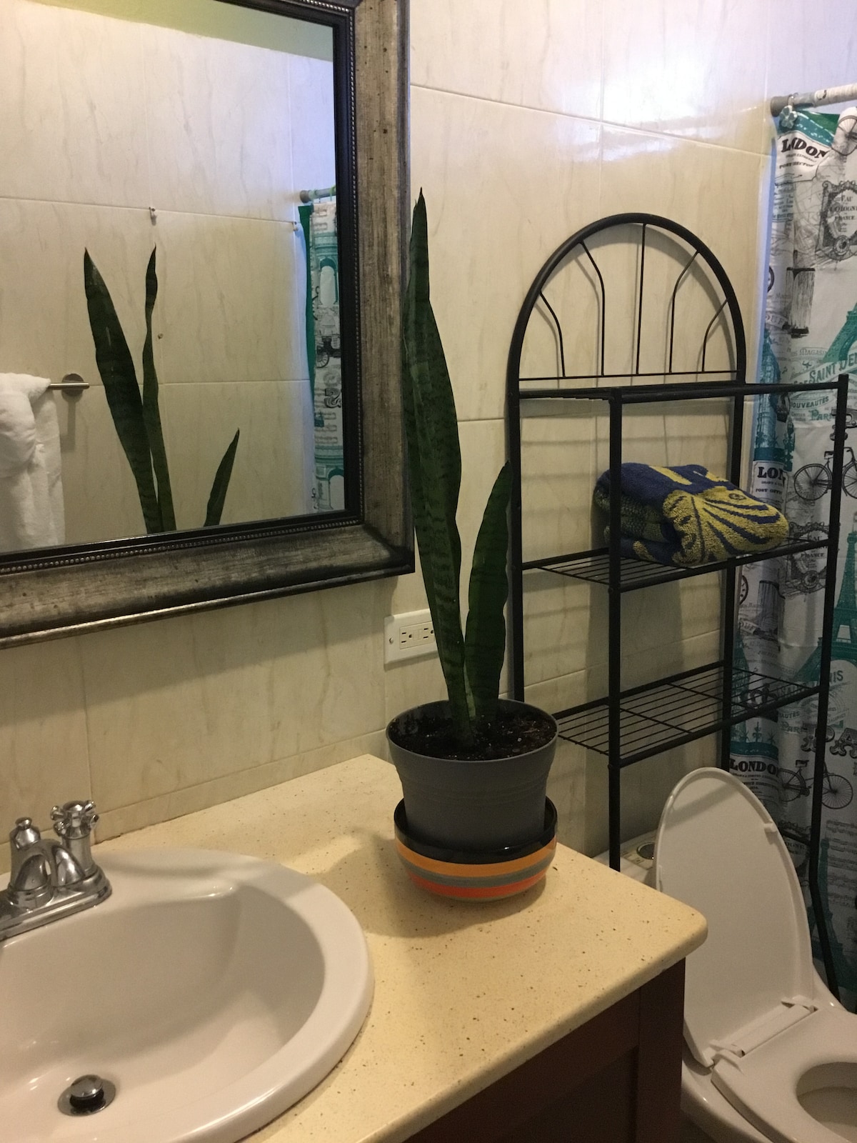 A bright bathroom features a large mirror reflecting a decorative plant and a towel rack. A plant pot sits beside a compact storage shelf, which holds a stack of towels. The sink area is clean and organized, enhancing the functionality of the space.