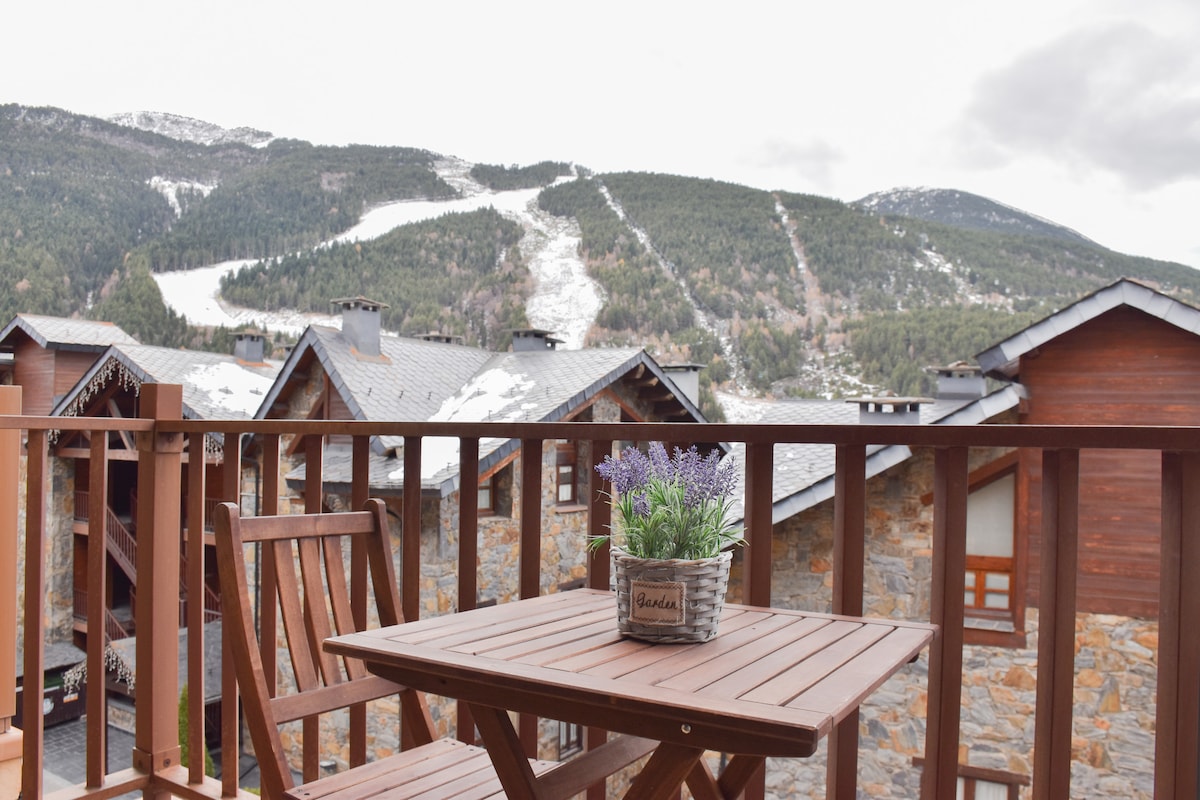 A wooden terrace table is adorned with a potted plant, offering a view of the surrounding mountains. Snow-covered slopes are visible in the distance, and rustic wooden buildings blend harmoniously with the natural landscape.