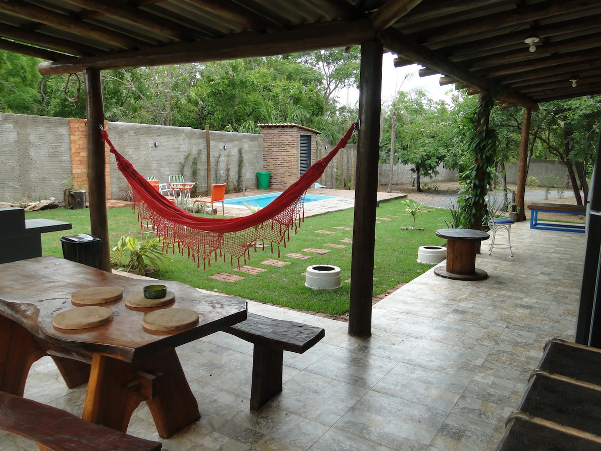 An outdoor entertainment area is visible, featuring a wooden dining table with several seats. A red hammock is draped between two posts. In the background, a private pool is surrounded by a grassy space and shaded by trees, creating a tranquil setting.