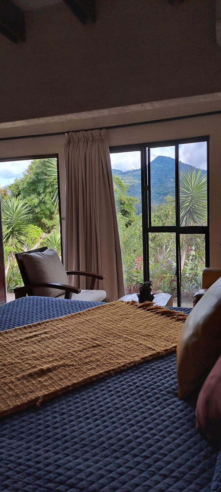 A cozy sleeping area is highlighted by a spacious bed adorned with a textured throw. Large windows provide views of the surrounding landscape, including mountains and lush foliage. A comfortable reading chair is positioned nearby, inviting relaxation within the serene environment.