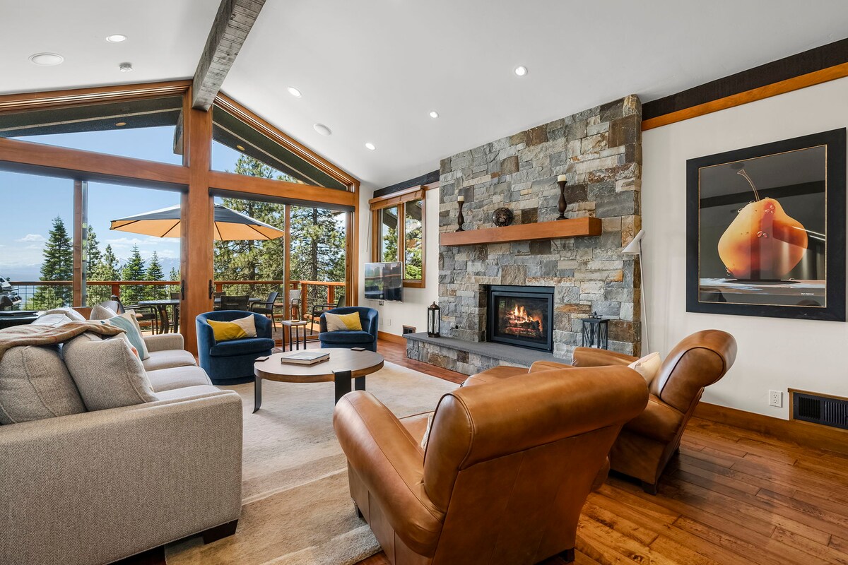 The living room features a stone fireplace framed by wood accents and large windows that allow natural light and lake views. Comfortable seating is offered by two brown leather chairs and a plush gray sofa. A round coffee table sits in the center of the space, enhancing the inviting atmosphere.