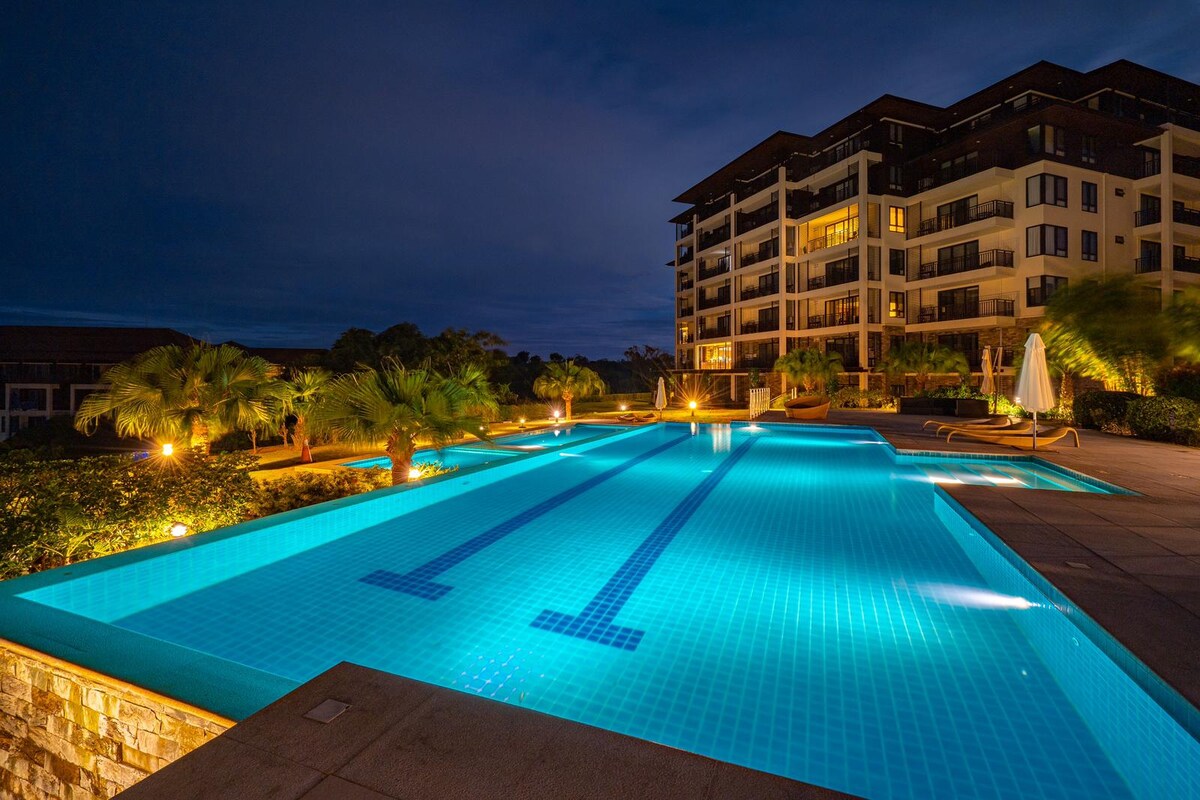 A large outdoor swimming pool is illuminated with soft, inviting lights, creating a tranquil atmosphere at night. Surrounded by lush landscaping, the pool features clear blue water and is framed by modern condominium buildings in the background.