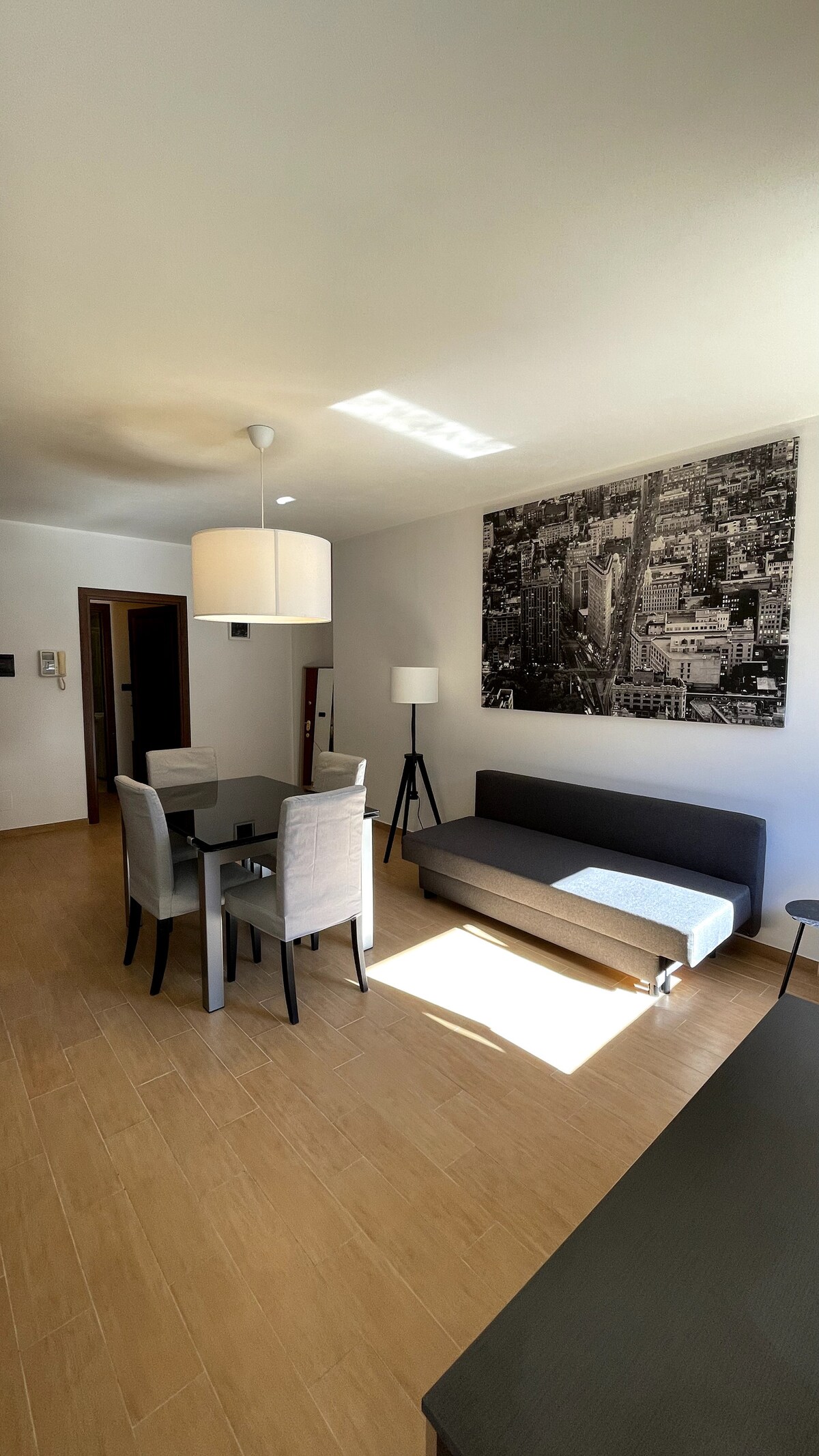 An open living area features a dining table with four chairs adjacent to a sofa. A large black and white cityscape photograph decorates the wall. Natural light enters from a nearby window, highlighting the warm wood flooring and modern furnishings.