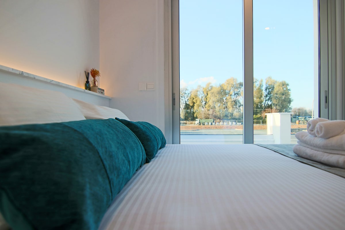 A spacious bed is dressed in white linens and accented by two turquoise pillows. Large glass doors provide natural light and an unobstructed view of greenery, creating a serene atmosphere. Neatly rolled towels are placed beside the bed.
