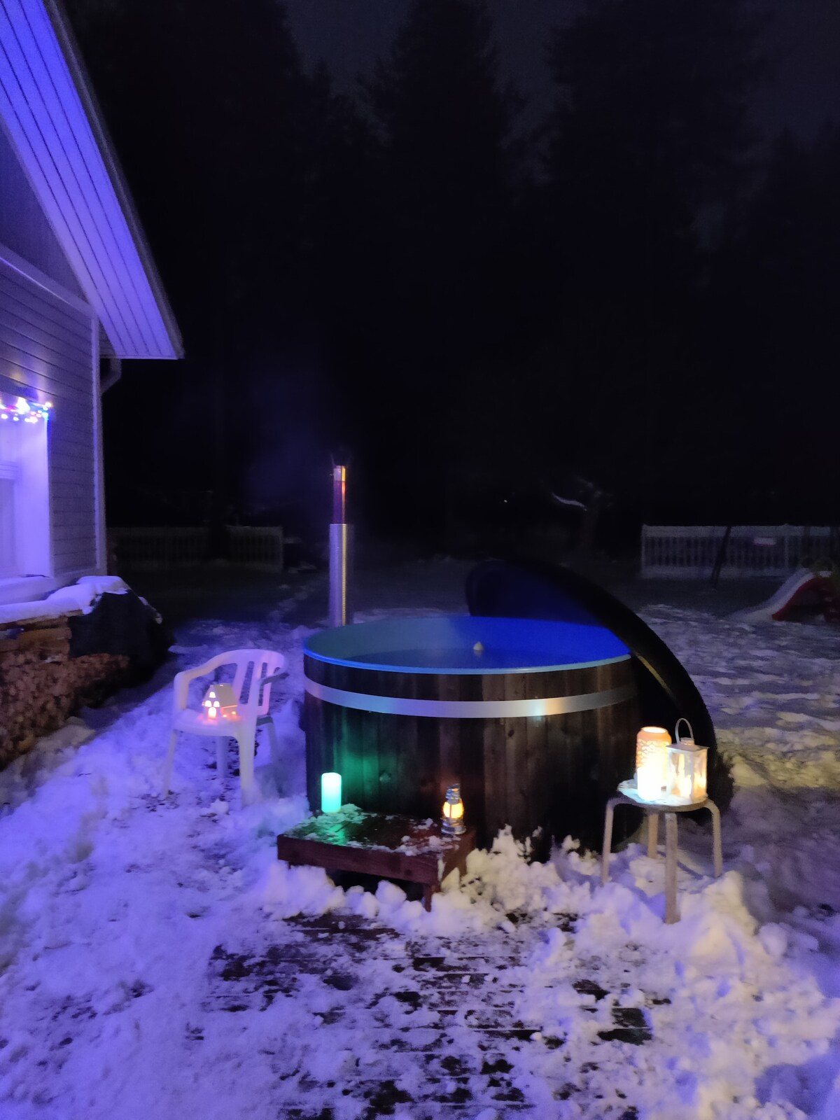 A hot tub is situated outdoors in a snowy landscape, illuminated by various lighting elements in soft hues. A simple wooden platform holds additional decorative lanterns, while white chairs provide seating nearby, creating a relaxing atmosphere in the winter setting.
