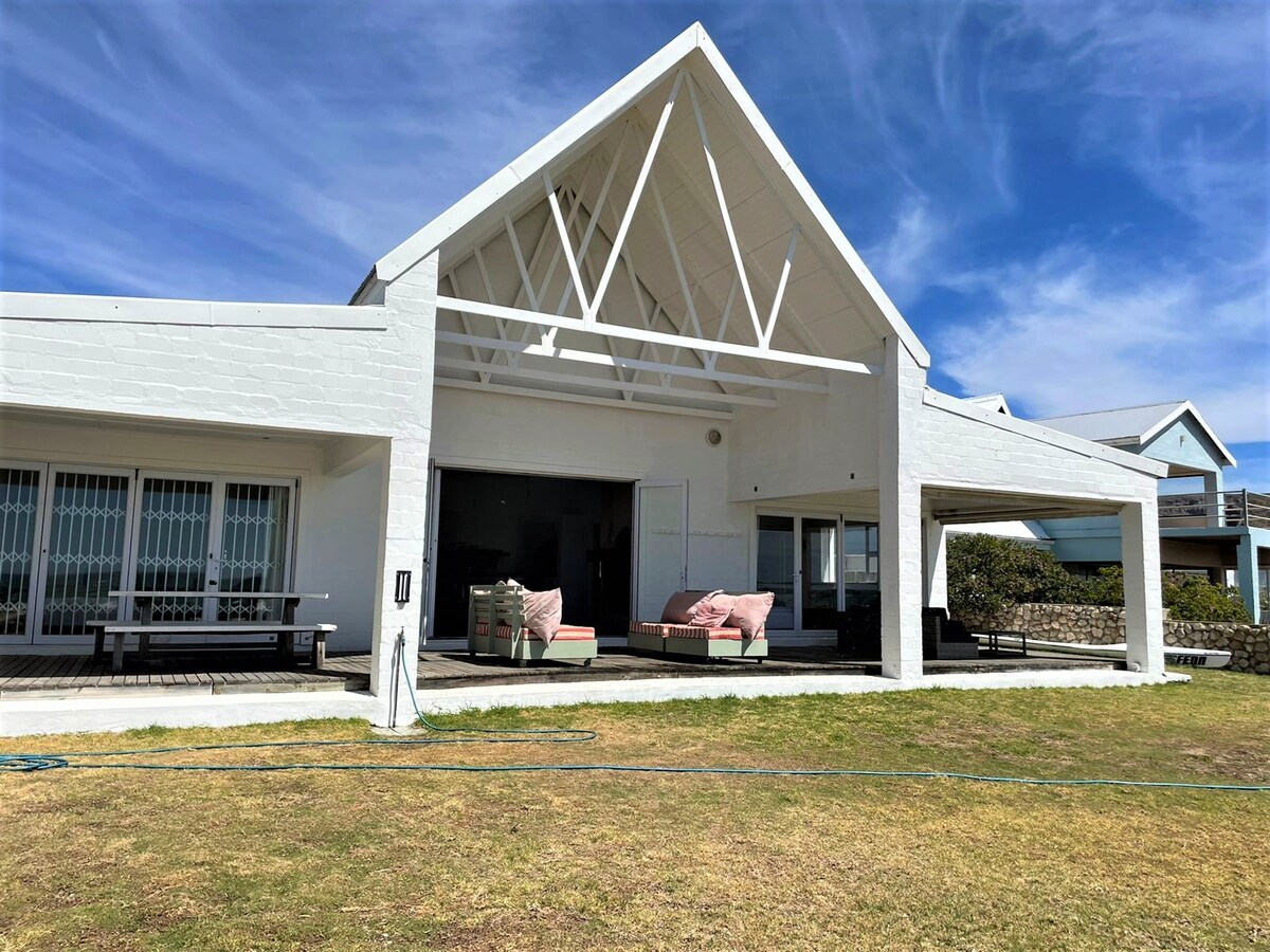 The spacious beach house features a high, light-filled ceiling and an open patio area. Comfortable seating options are provided, alongside a well-maintained garden and views of the sky. The structure exhibits a white exterior, blending with the surrounding landscape.