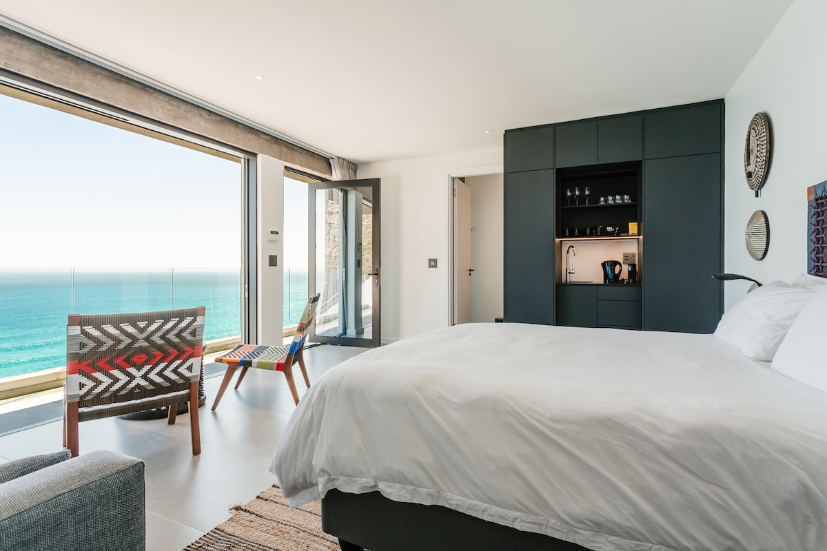 A modern studio showcases an ocean-facing queen bed with crisp white bedding. A spacious terrace offers stunning sea views through large sliding doors. Lounge seating is visible, complemented by a compact kitchenette area featuring coffee-making amenities. Natural light fills the room, enhancing the sleek design.