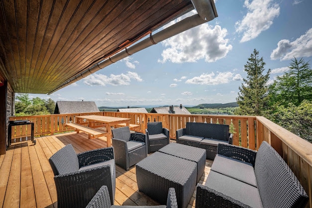 A spacious wooden terrace is seen, featuring a large outdoor dining table and several dark wicker lounge chairs. The scenic view includes open skies and distant rolling hills, complemented by a barbecue grill at one corner.