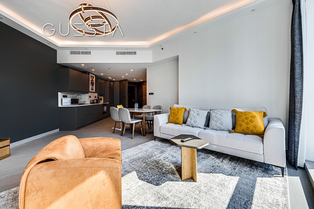 A contemporary living area is highlighted by a plush sofa adorned with patterned cushions and a cozy armchair. A sleek wooden coffee table sits on a soft area rug, with the kitchen and dining area visible in the background, featuring modern furnishings and ample natural light.