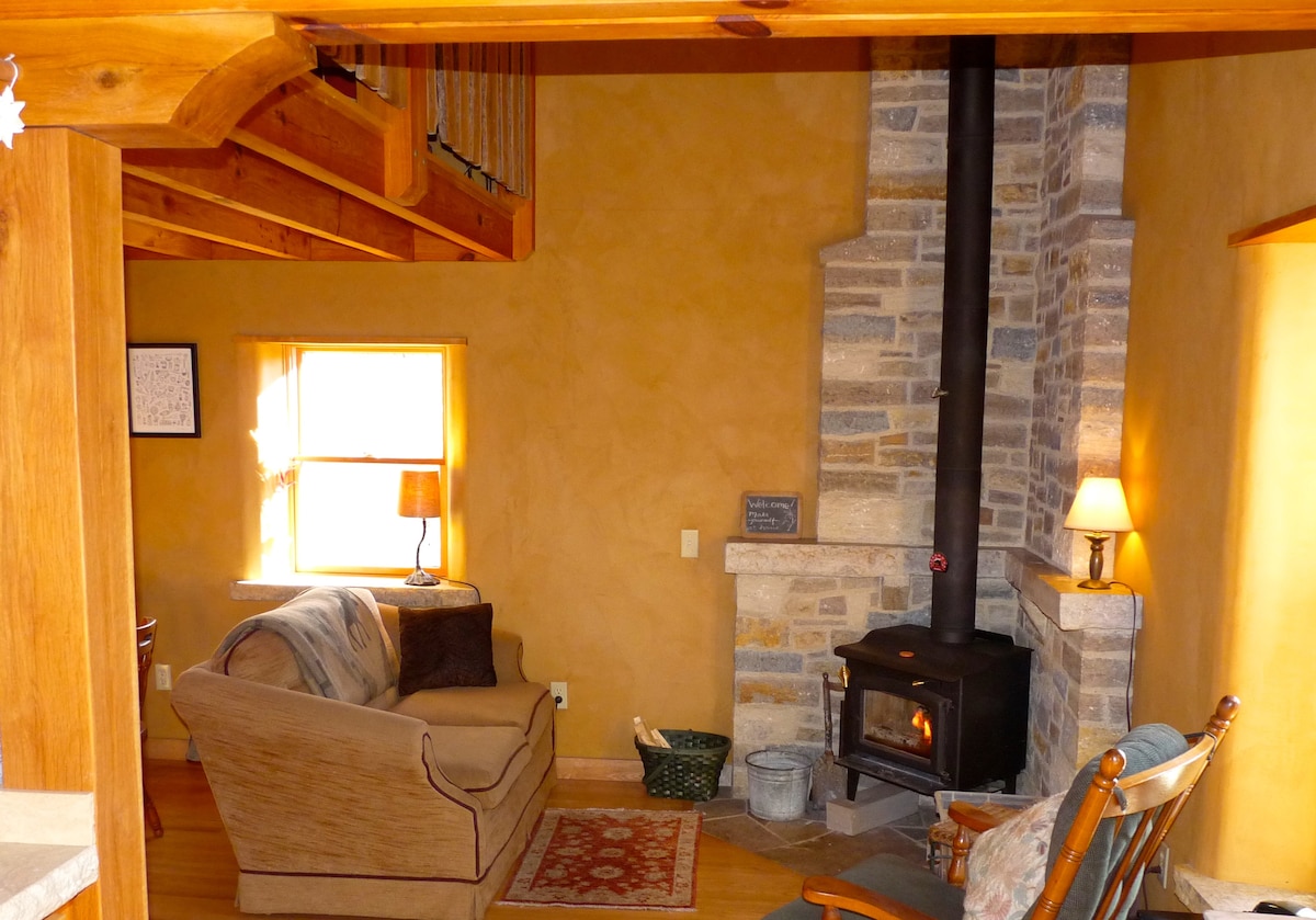 An inviting living area features a cozy couch facing a wood-burning stove encased in local stone. Warm light from nearby lamps creates a comfortable ambiance. Exposed wooden beams are visible in the ceiling, and a soft rug adds a touch of warmth to the oak floor.