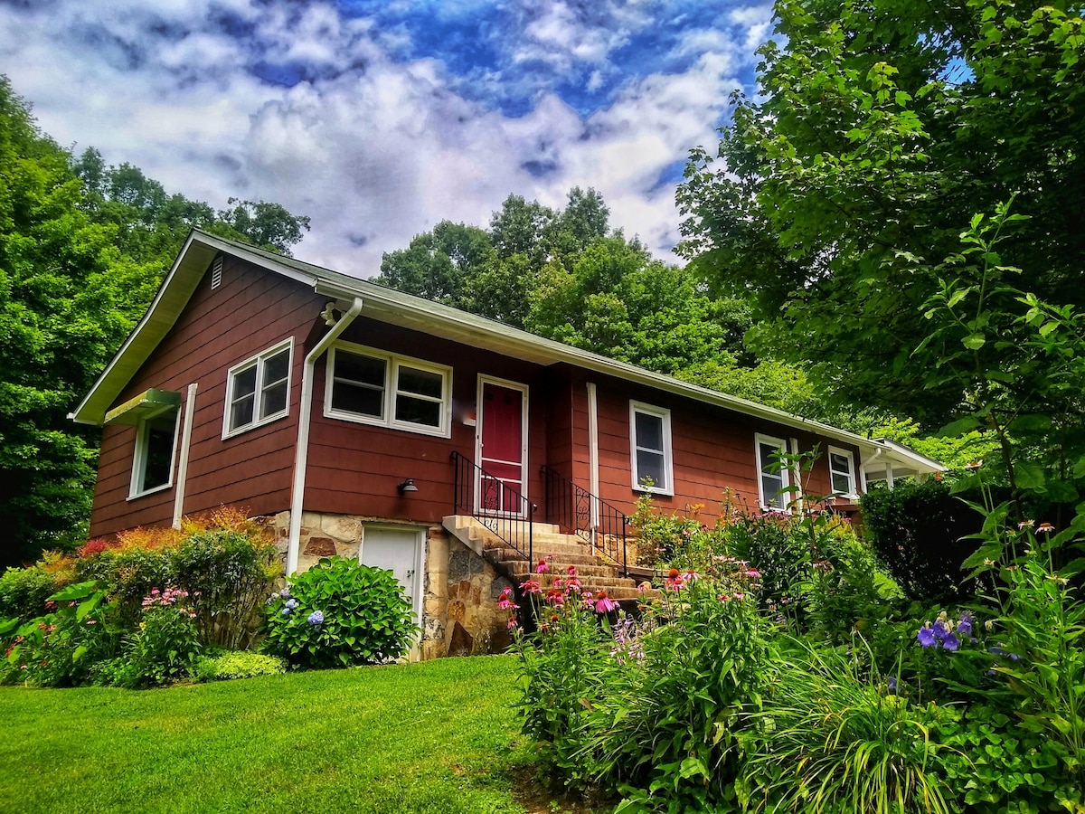 The cottage features a charming exterior with red siding and a welcoming front door. Lush greenery surrounds the house, complemented by vibrant flower beds. The gently sloped lawn leads to a stone foundation, enhancing the home’s rustic appeal against a backdrop of trees and a cloudy sky.
