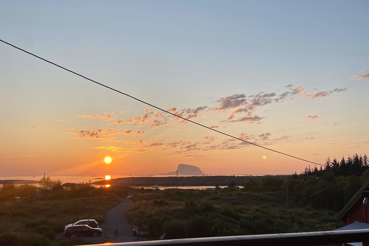 A sunset view over the water is captured, featuring hues of orange and yellow in the sky. Silhouetted hills and trees are visible in the background, with a distant rock formation adding depth to the landscape. A vehicle is parked along a gravel path in the foreground.